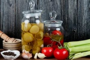 Side view of jars of pickled tomato with vegetables black pepper and garlic crusher on wooden background with copy space