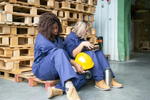 Tired sleepy diverse female plant workers sitting on wooden pallets with thermos and cookies