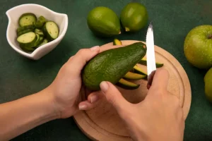 Top view of female hands cutting a bright green avocado with knife on a wooden kitchen board with limes and green apples isolated on a green wall